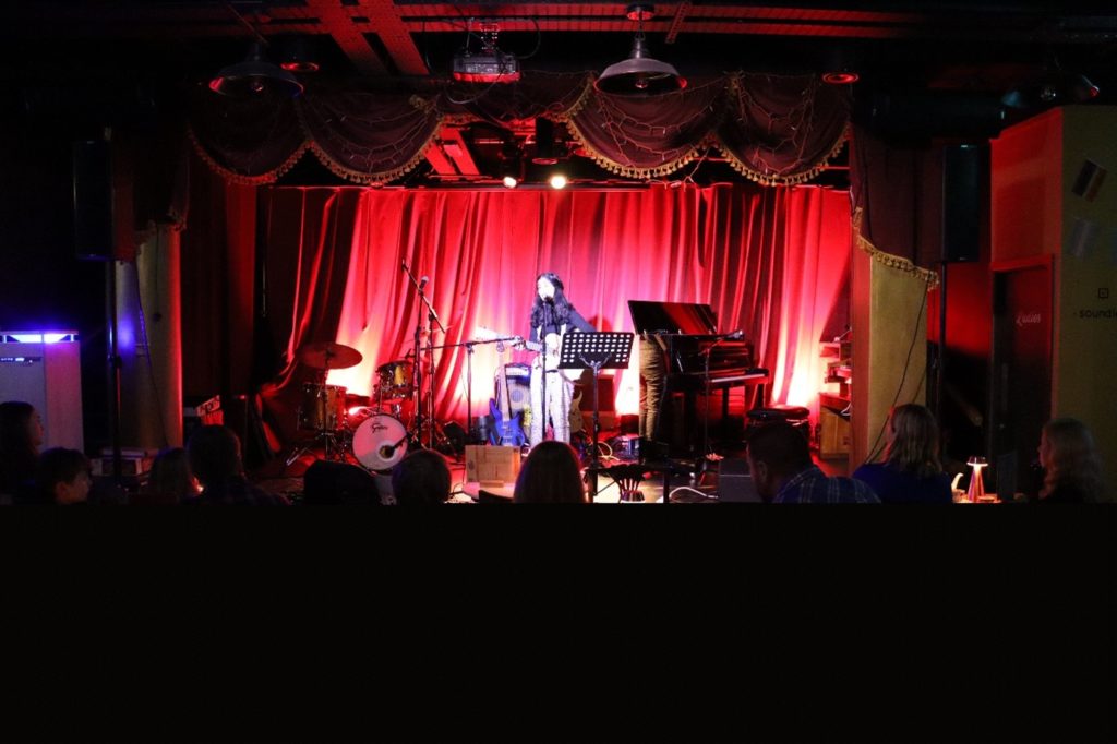 A colour photo of a young guitarist performing on a stage with lighting and a red curtain