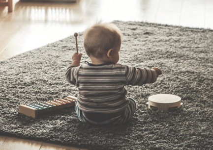 A small child/ baby sits on the floor playing with a drum and xylophone.