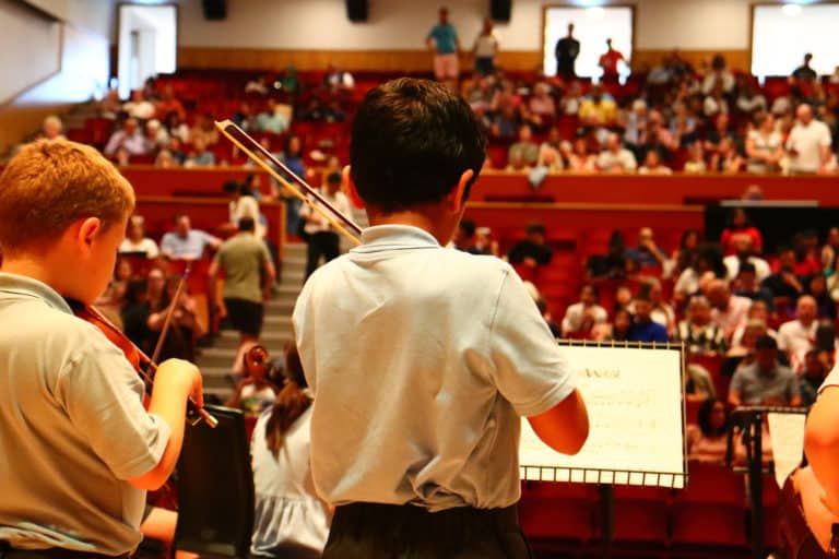 A photo of a student playing violin. The student's back is facing the camera and we can see the audience taking their seats in the venue.