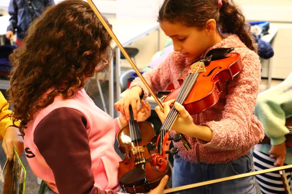 Two young girls dressed in pink clothing, one is helping the other with finding a note on the violin