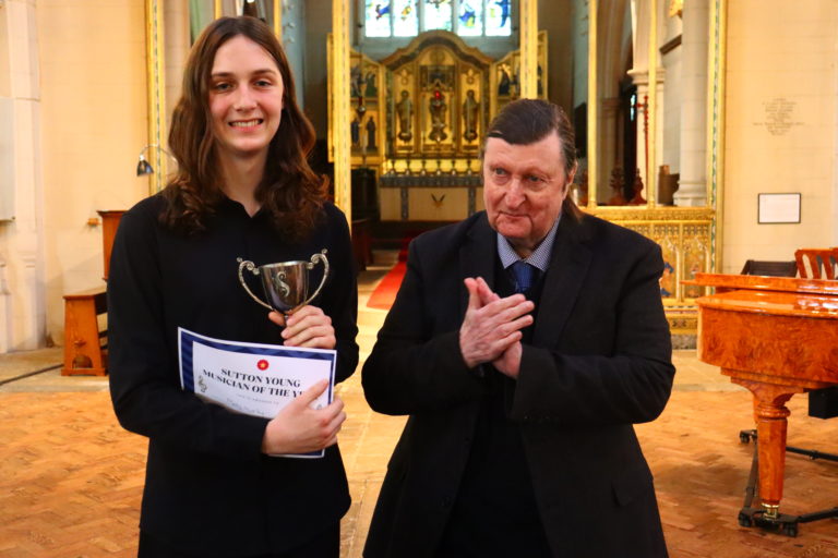 A smiling student dressed in black with a trophy and certificate in a church. The adult next to the student is smiling and clapping