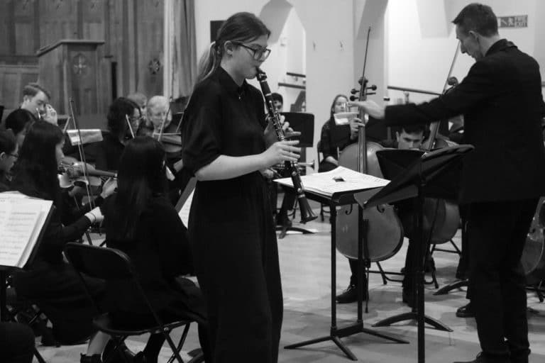 A black and white photo of a girl playing clarinet in front of an orchestra in a church