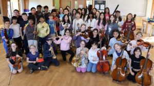 A group of children with instruments smiling, in a hall