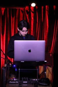 A young DJ performing with a silver laptop in front of a red curtain