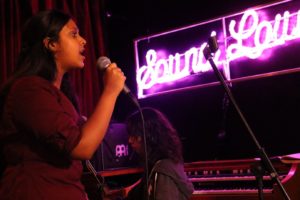 A picture of a student singing and and another playing piano. A pink neon light behind them saying "Sound Lounge"