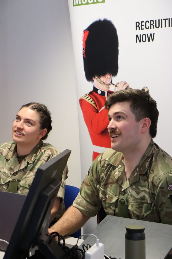 2 people in military clothes sit in front of a British Army Music banner talking to a family who is off screen