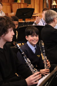 Two young students holding their clarinets in a band rehearsal. One of them, a younger boy with dark brown hair is smiling at the camera.